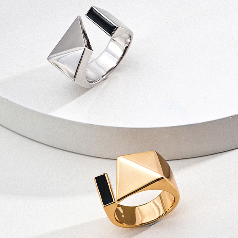 Side-by-side view of silver and gold variants of the S925 open ring, each featuring a geometric pyramid on one end and a black enamel rectangular inset on the other, arranged on layered ceramic surfaces.