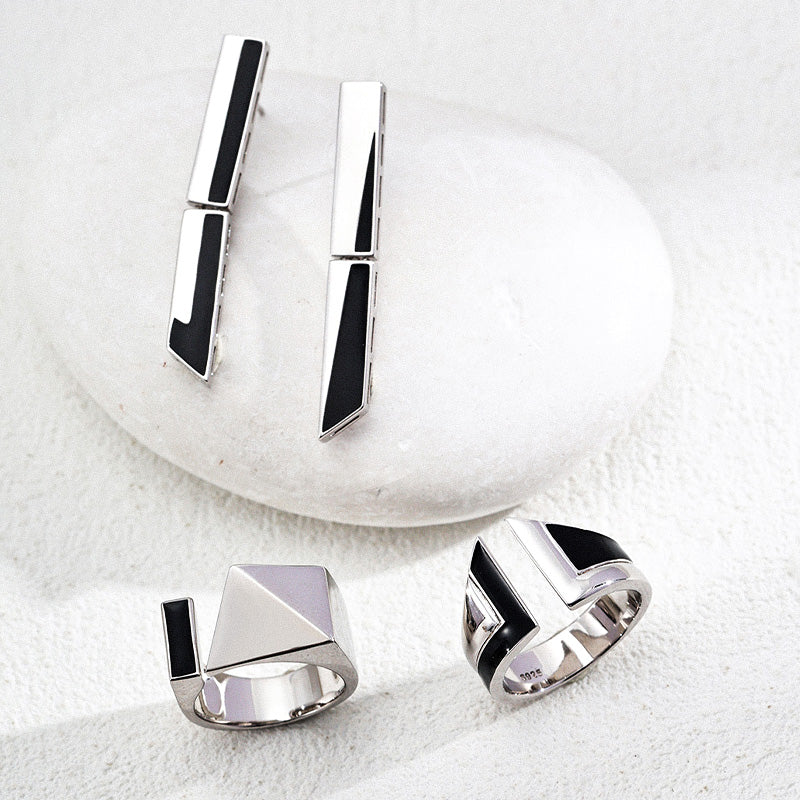 A contemporary jewelry trio featuring silver-tone S925 earrings with architectural black enamel detailing, paired with two matching geometric rings—one with a bold pyramid structure and the other with clean segmented curves—styled on a smooth stone surface for a sleek, modern look.