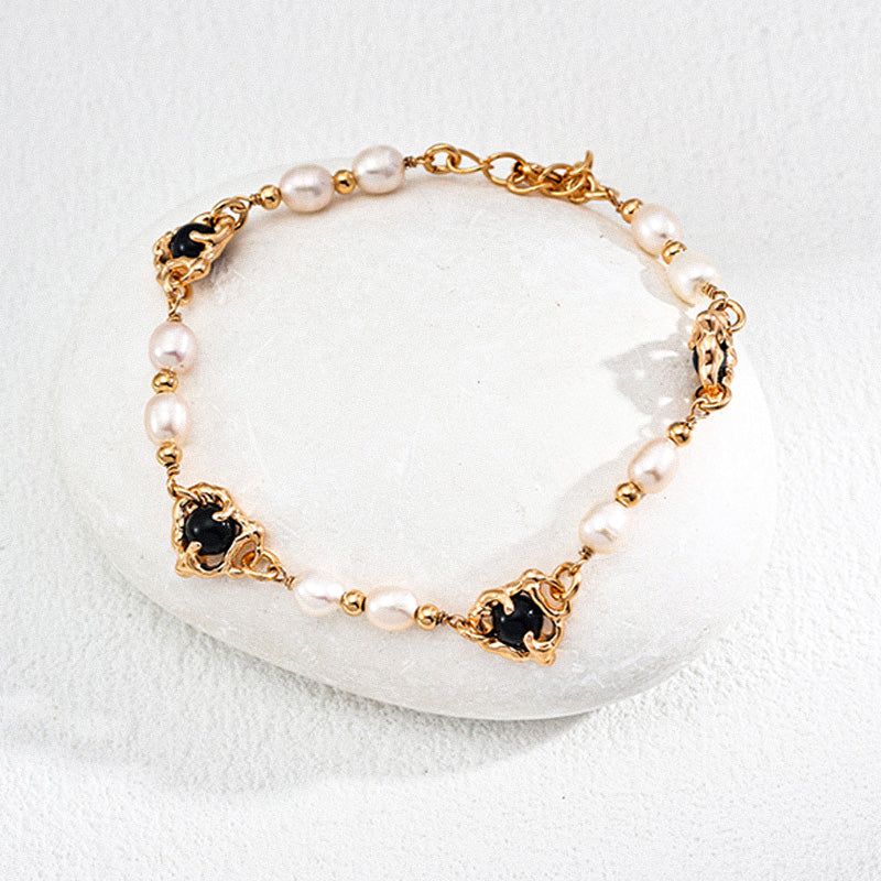A close-up of a vintage-style bracelet crafted in S925 silver, featuring alternating natural pearls and black onyx stones with ornate gold-toned floral settings, laid on a smooth white stone.