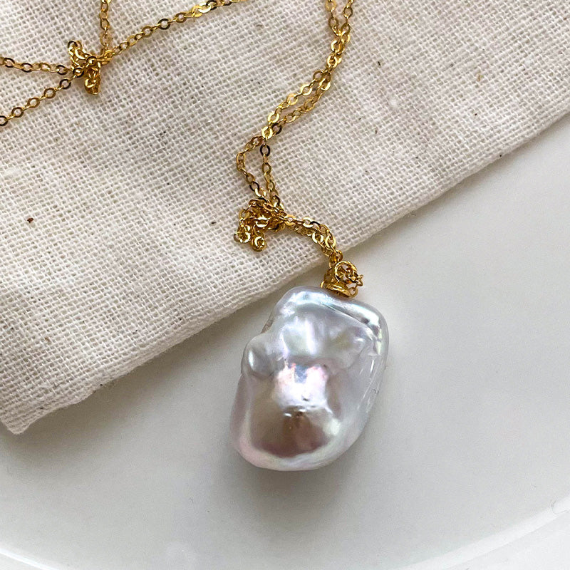 White baroque pearl pendant necklace with 14K gold chain resting on linen fabric.