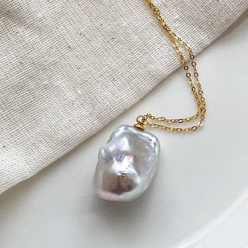 Lustrous baroque pearl pendant in soft white, paired with a 14K gold chain, displayed on neutral linen.