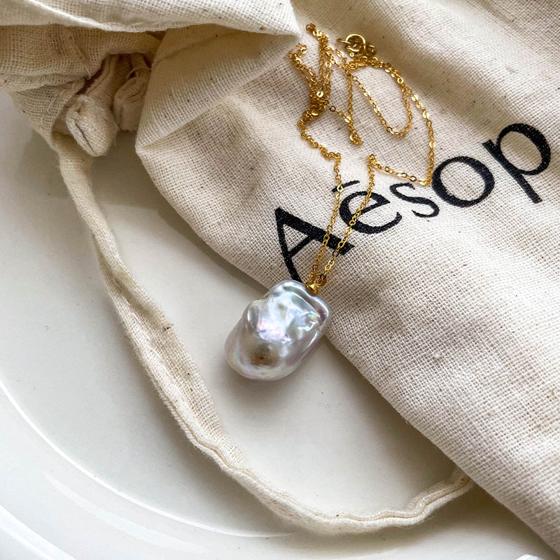 Elegant white baroque pearl pendant necklace with 14K gold chain, styled on an Aesop drawstring pouch.