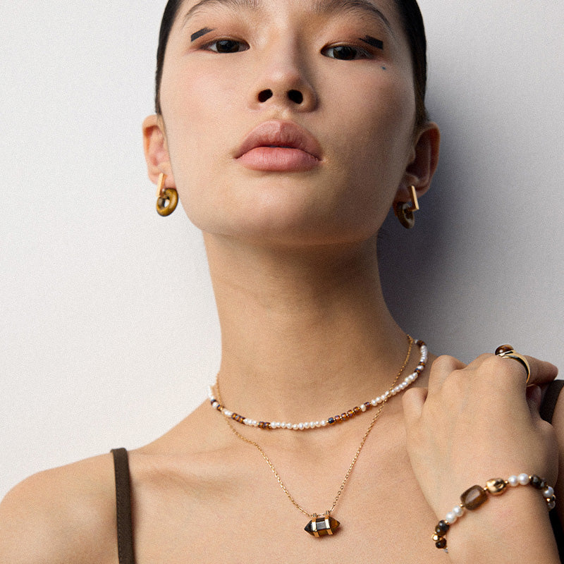 A model wears the S925 sterling silver geometric tiger’s eye pendant necklace layered with a pearl beaded choker and matching bracelet, showcasing a warm, earthy-toned jewelry styling.
