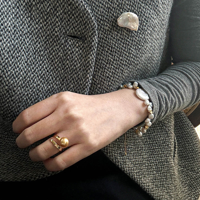 Elegant styling of a baroque pearl and gold bracelet on a woman's wrist, paired with a statement pearl ring and textured grey outfit.