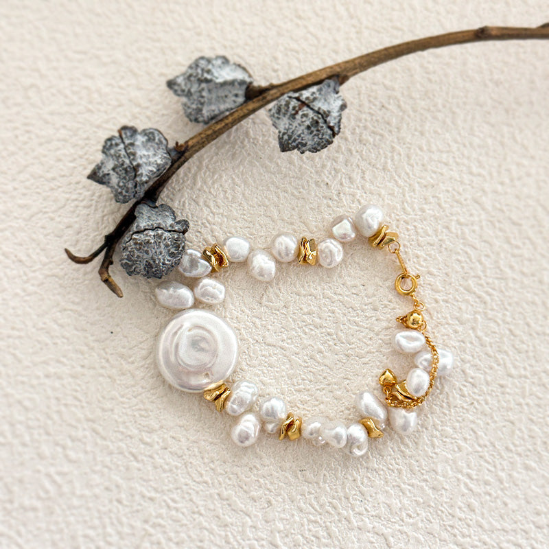 Styled composition of a baroque pearl bracelet with gold details, arranged beside a dried botanical branch on textured ivory background.